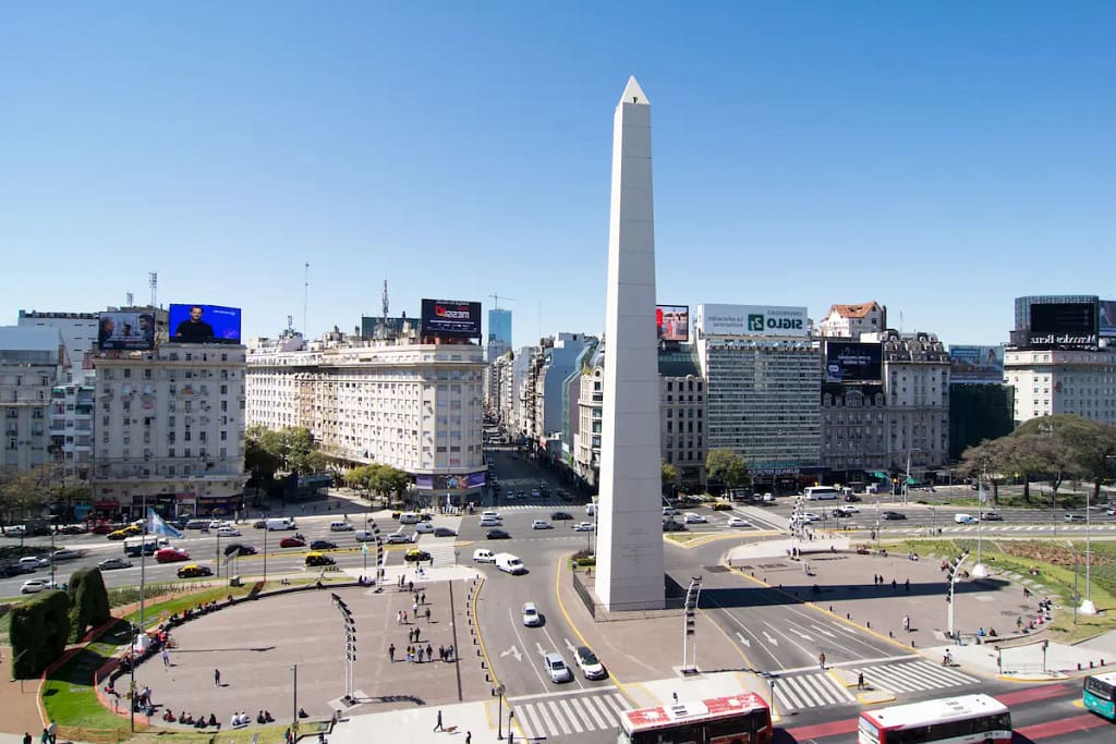 Una escena vibrante que captura el icónico Obelisco erguido en medio del bullicioso tráfico de la Avenida 9 de Julio en Buenos Aires, Argentina. Esta fotografía encapsula la energía de la ciudad, mostrando su rica arquitectura y estilo de vida urbano