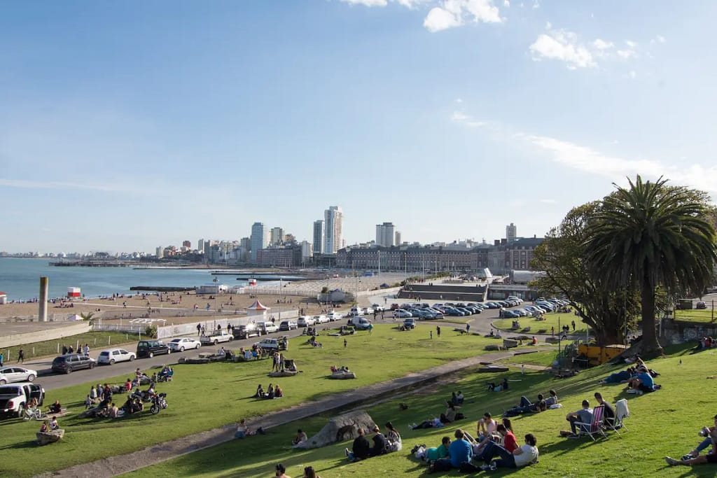 Una vista impresionante del paisaje urbano de Mar del Plata, Provincia de Buenos Aires, Argentina, al atardecer, con colores vibrantes en el cielo. Los turistas disfrutan de su tiempo sobre la hierba de la playa, creando una atmósfera animada.