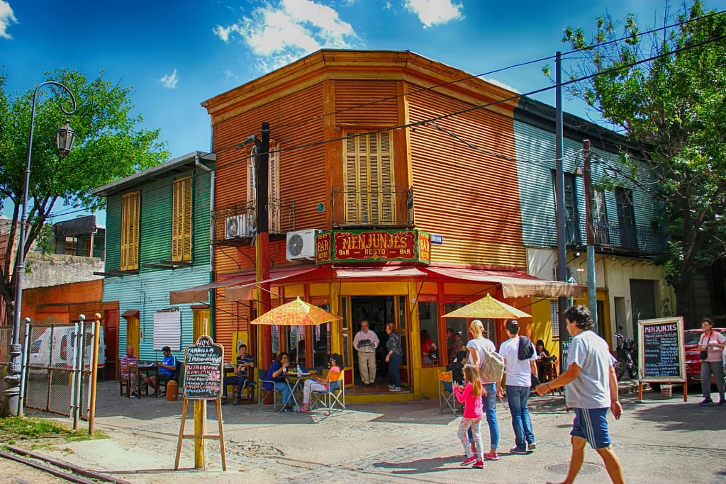 Turistas caminan por calle empedrada frente a casas de madera en barrio La Boca, Buenos Aires, Argentina.