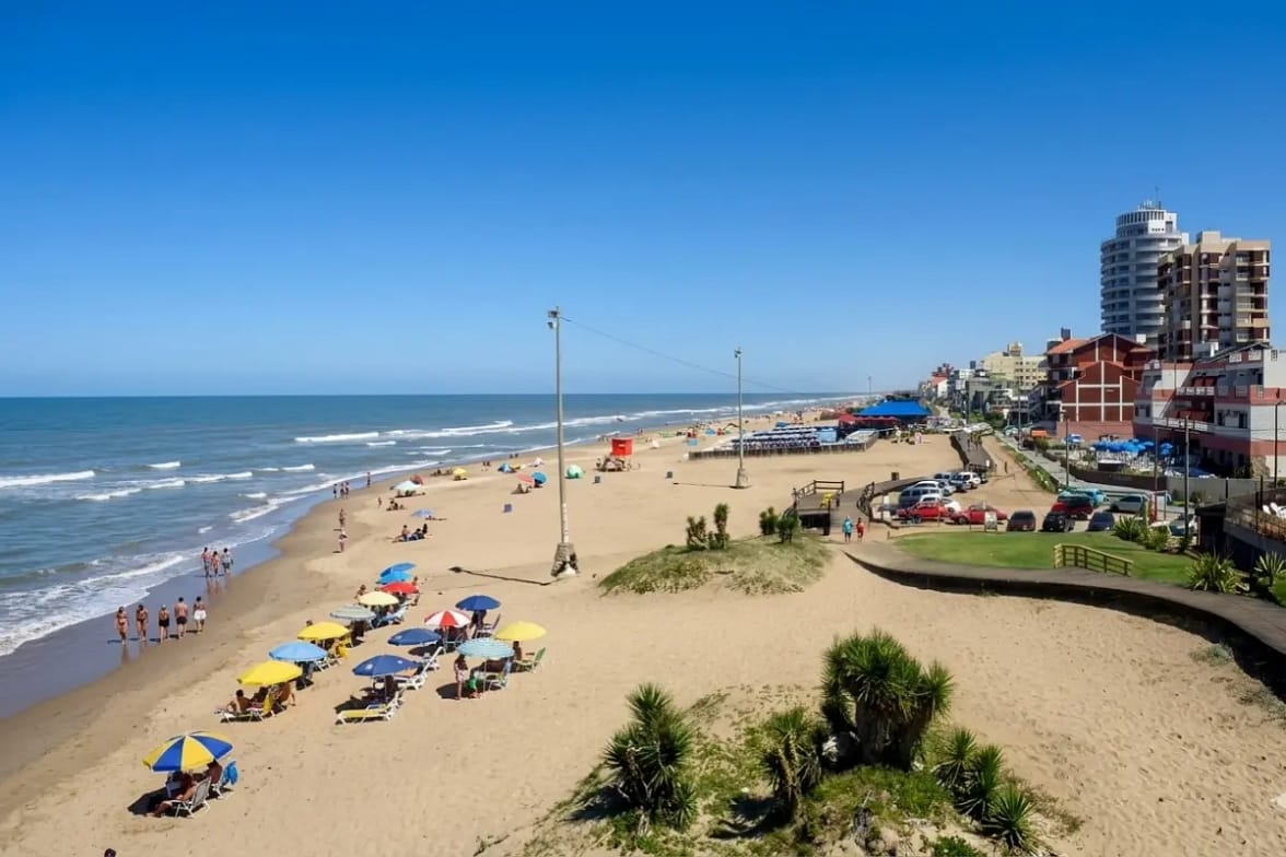 Villa Gesell, Región Buenos Aires, Argentina: Verano en la Costa Atlántica Una toma aérea oblicua de gran angular que captura la extensa playa de Villa Gesell, Buenos Aires, en pleno verano. Miles de personas disfrutan del sol bajo sombrillas de colores, paseando por la orilla y bañándose en el mar Atlántico turquesa con olas suaves, bajo un cielo azul brillante con nubes dispersas.